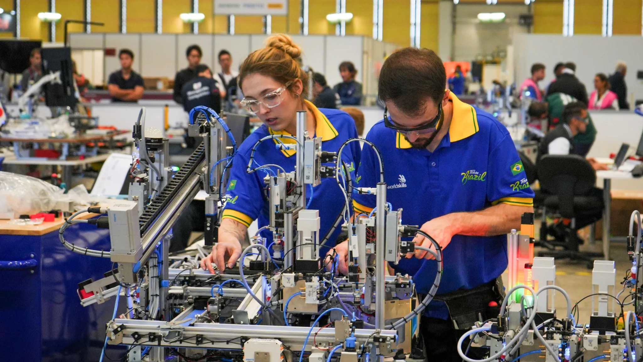 Dois jovens, um homem e uma mulher, estão trabalhando juntos em um projeto que envolve equipamentos eletrônicos e fios. O homem está de costas para a câmera e veste uma camiseta azul com um logotipo amarelo. A mulher está de frente e veste uma camiseta amarela com o mesmo logotipo. Eles parecem estar engajados em uma atividade prática do setor industrial.